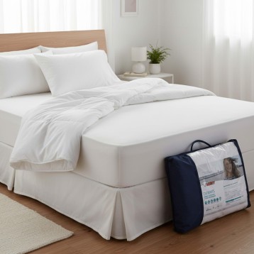 Cama con cabecero de madera clara y ropa de cama blanca junto a protector de colchón en bolsa azul marino