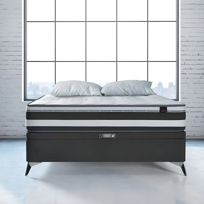 Cama con base tapizada negra y patas metálicas frente a ventanal con vistas urbanas y dos almohadas grises