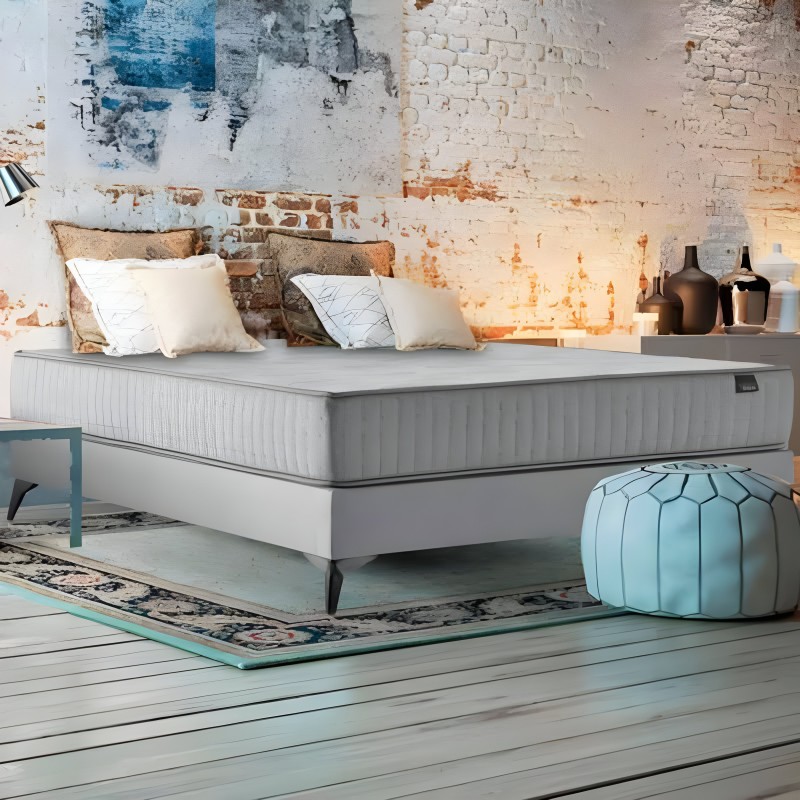 Cama con base blanca y colchón gris tapizado con almohadas decorativas en ambiente industrial con pared de ladrillo visto